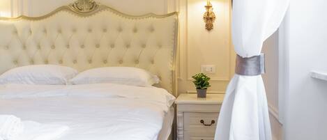 A neatly made bed with white linens, a tufted headboard, a bedside table with a lamp, and a small potted plant.