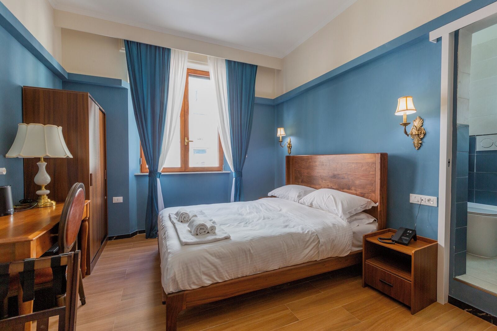 A bedroom with a wooden bed, a desk with a lamp, and a bathroom visible through a glass door.