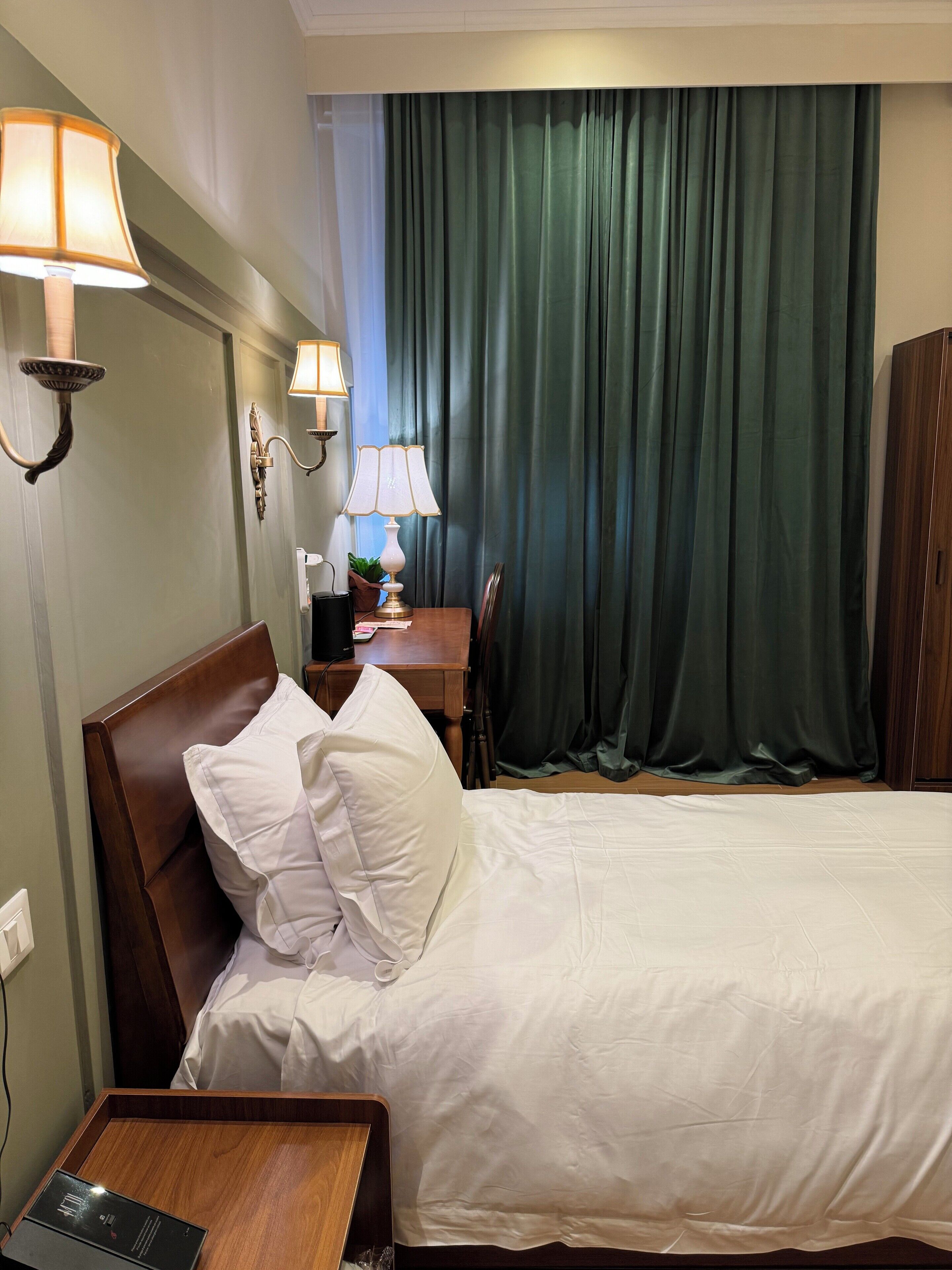 Quarto de hotel com cama, mesas de cabeceira, luminárias, escrivaninha e cortina.