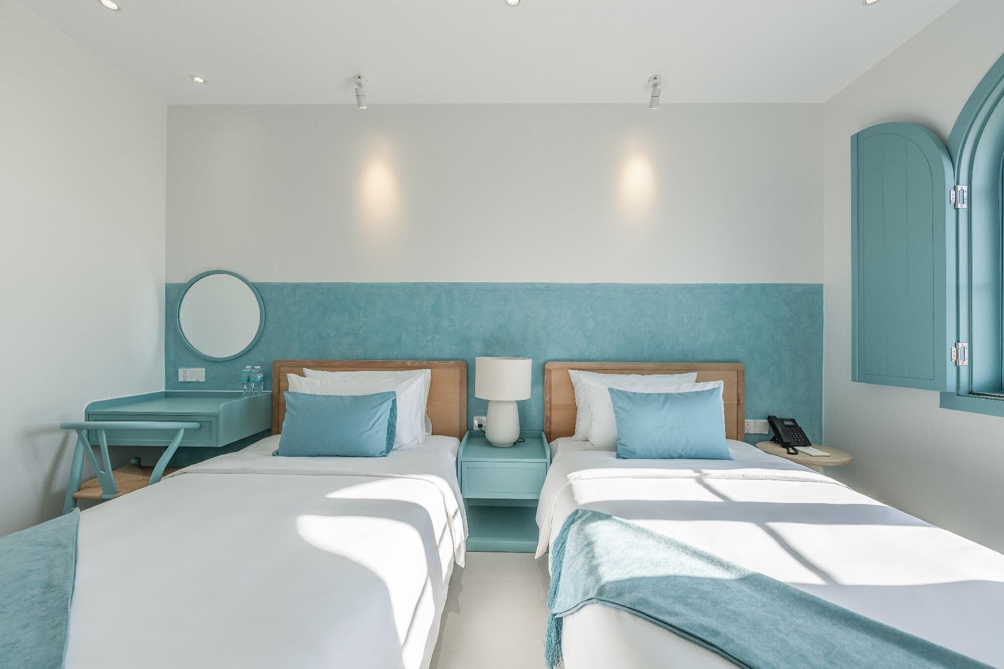 A modern hotel room with two beds, a desk, a round mirror, and a nightstand with a phone.
