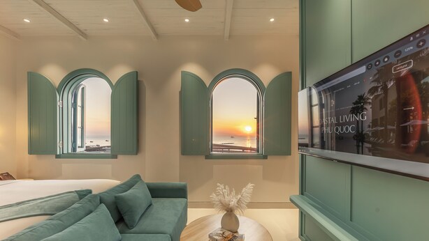 A modern hotel room with a bed, teal-colored sofa, a round wooden table with a vase, and two arched windows with a sunset view.