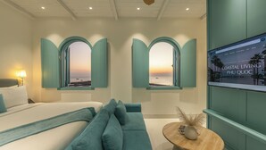A hotel room with a bed, a teal sofa, a round wooden side table, and two arched windows with a sunset view.