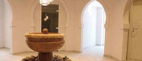A stone fountain with a wooden bowl in a hallway with arched ceilings and a door in the background.