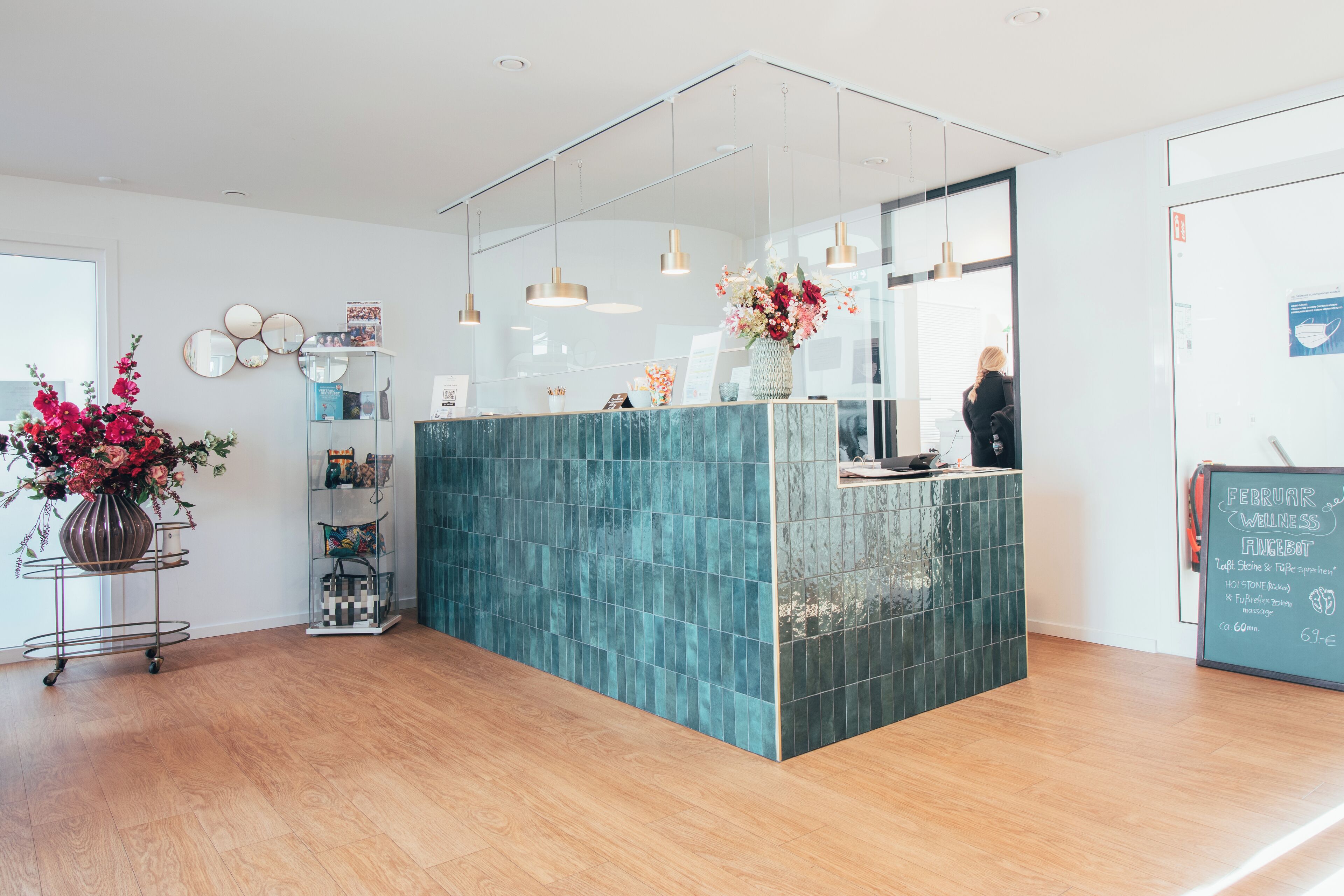 Un espace d’accueil doté d’un comptoir aux carreaux turquoise, d’un éclairage suspendu, d’un vase avec des fleurs, d’une étagère contenant divers objets et d’un tableau noir avec du texte.