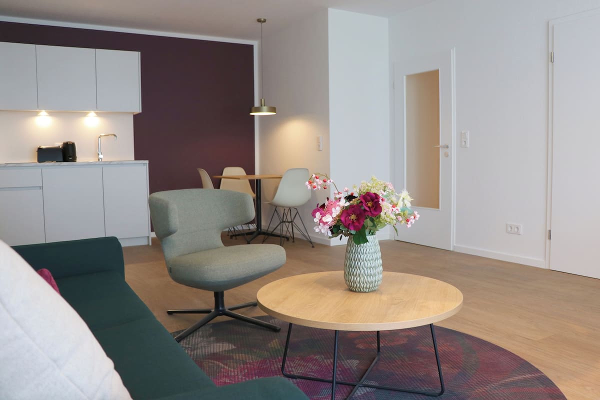 A modern living room with a green sofa, a round coffee table with a vase of flowers, a dining area with chairs, and a kitchen with white cabinets and a pendant light.