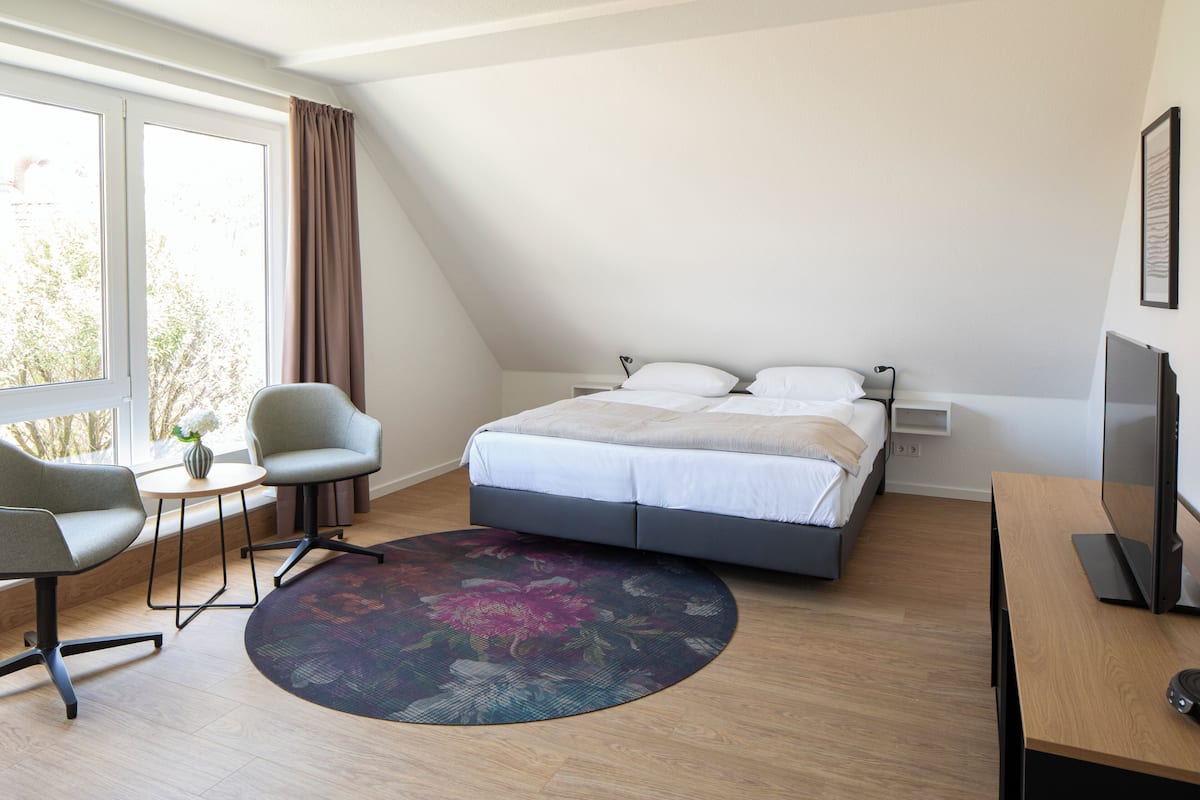 A modern hotel room with a bed, a desk, a chair, a small table, and a large window.
