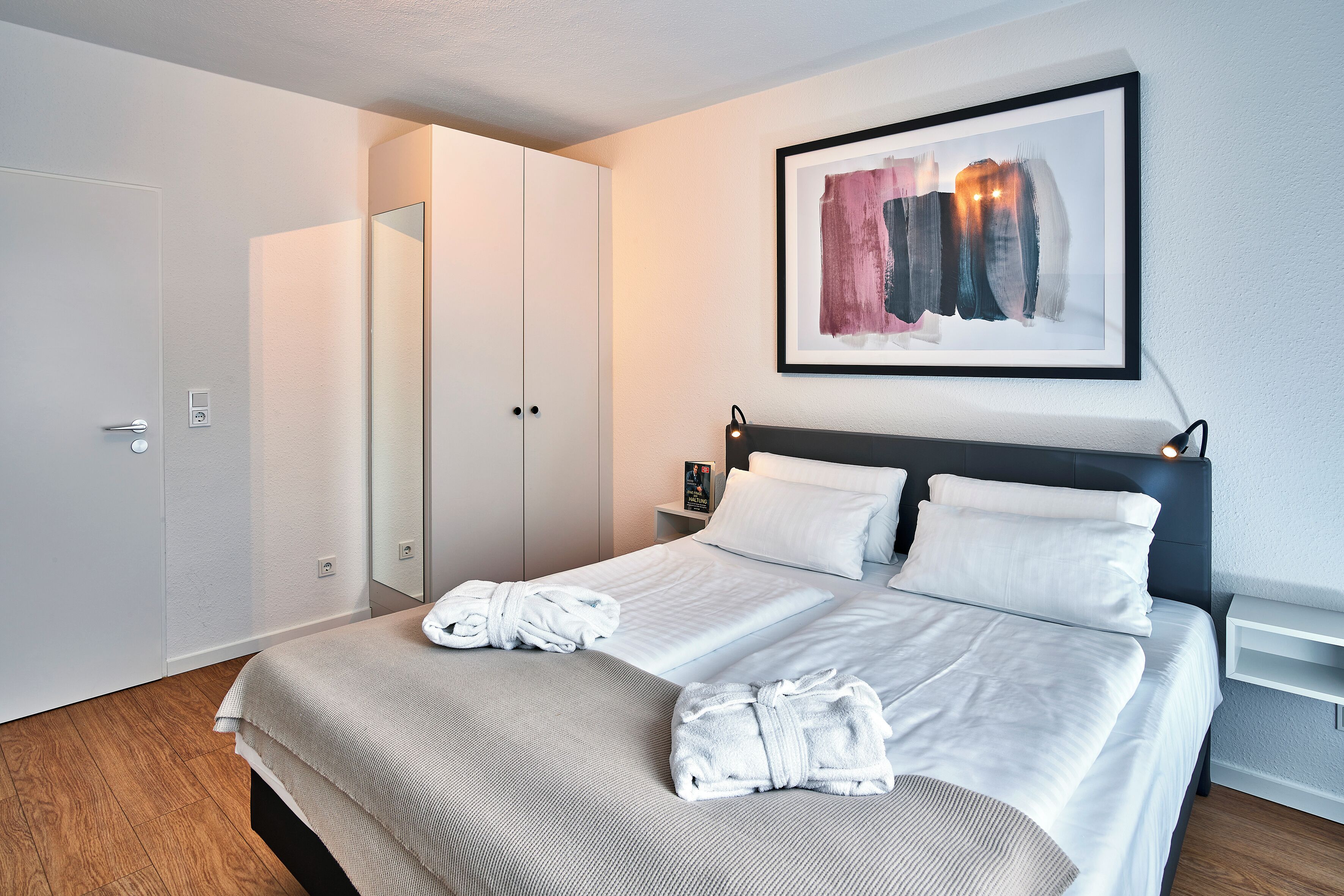A hotel room with a bed, two white robes, a framed abstract painting, a nightstand, and a wardrobe.