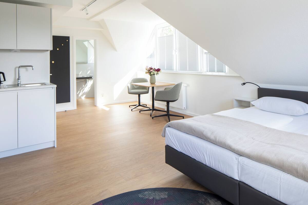 A modern hotel room with a large bed, a small desk, and a kitchenette.