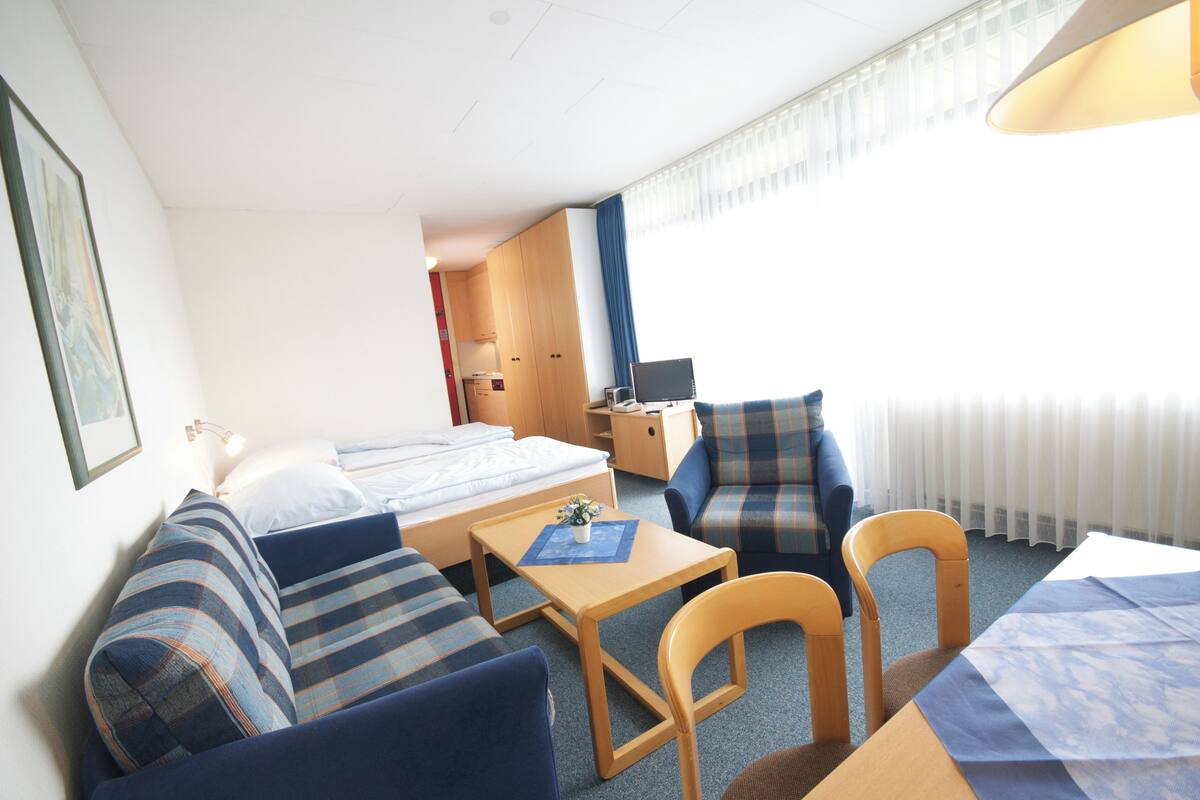 Ein Hotelzimmer mit Bett, Sofa, Schreibtisch und Sessel. Ein Fenster mit Vorhängen, eine Lampe und ein Bild an der Wand sind ebenfalls zu sehen.