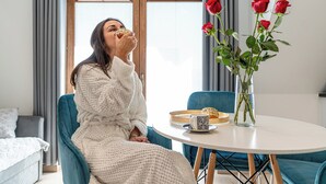 Eine Person sitzt an einem runden Tisch mit einer Rosenvase, einer Tasse und einem Gebäckteller.