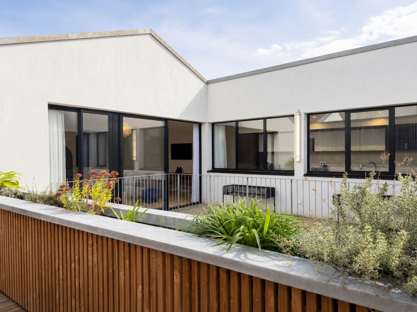Un edificio residencial moderno con amplios ventanales, un balcón con baranda de madera y un jardín con diversas plantas.