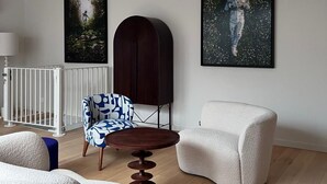 Ein modernes Wohnzimmer mit einem weiß-blauen gemusterten Sessel, einem runden Holztisch, einem weißen Sessel, einer dunklen Holzkommode und zwei gerahmten Kunstwerken an der Wand.