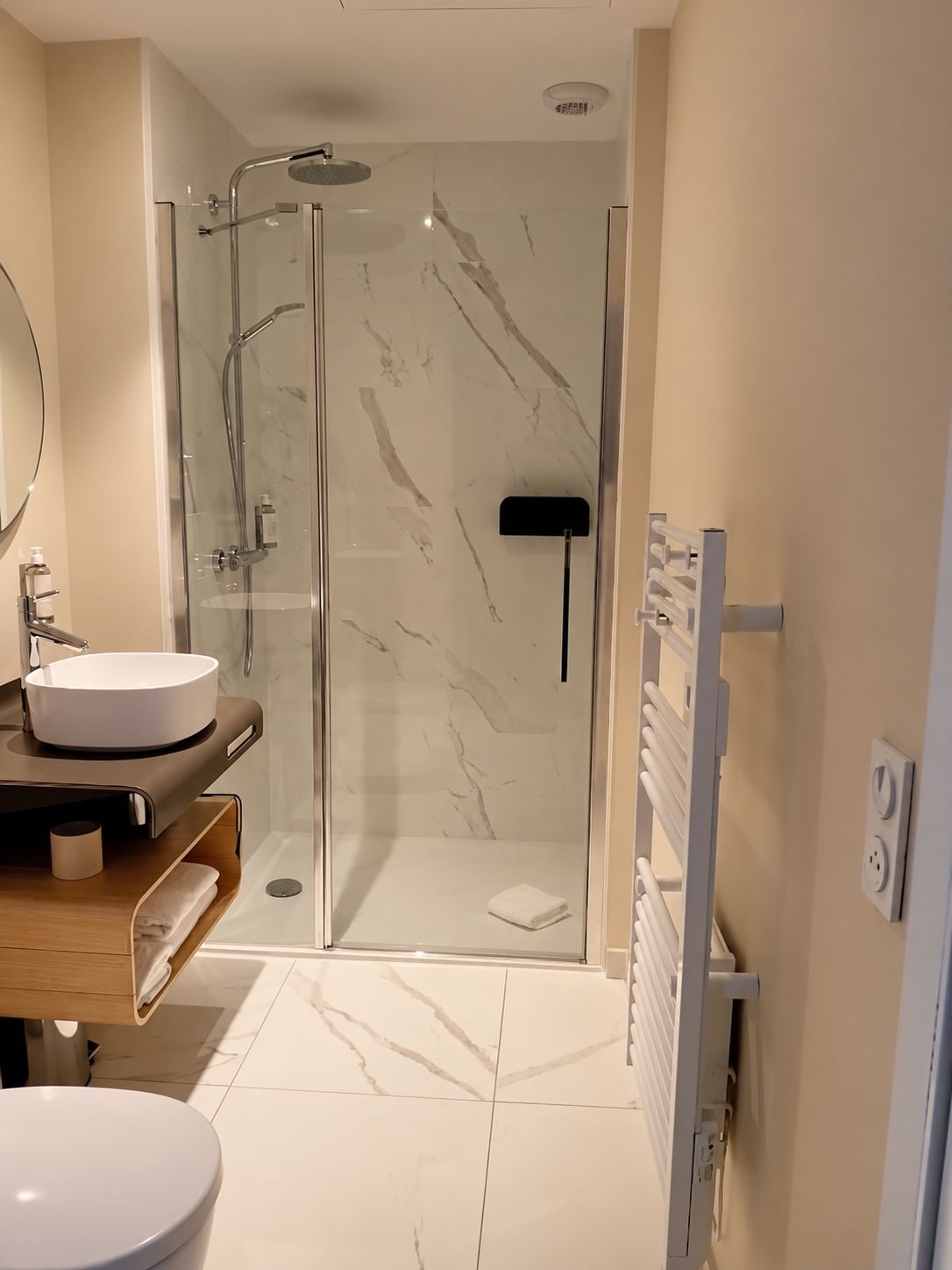 Een moderne badkamer met een douchecabine van glas, een rond wastafeltje en een handdoekrek.