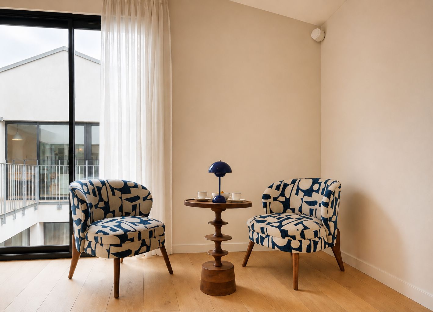 Une pièce avec deux fauteuils à motifs, une petite table avec une lampe et une vue sur un balcon.