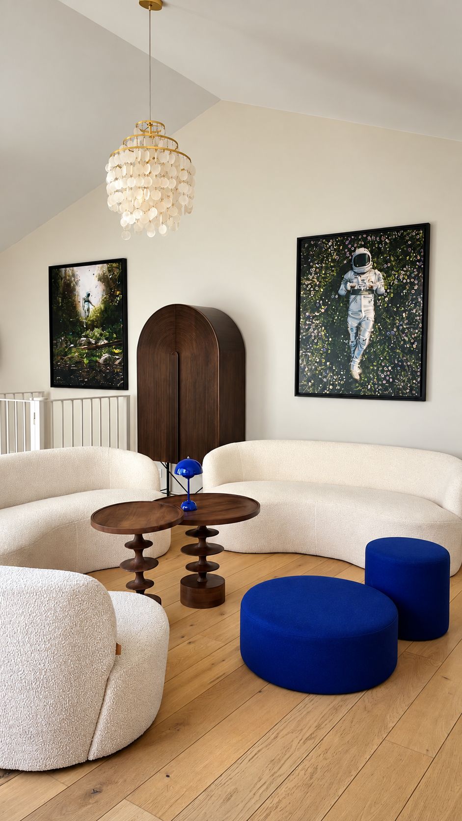 Een moderne woonkamer met een gebogen witte bank, een ronde houten salontafel en blauwe poefs. Twee ingelijste kunstwerken hangen aan de muur, een kroonluchter en een houten kast.