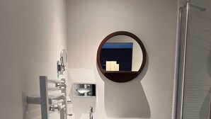 Un baño moderno con un tocador blanco, un espejo redondo, cerramiento de ducha de vidrio y un mueble de madera oscura.