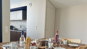 Ein Essbereich mit einem Holztisch, gedeckt für vier Personen, umgeben von Korbstühlen mit blauen Kissen. Im Hintergrund ist eine Küche erkennbar.