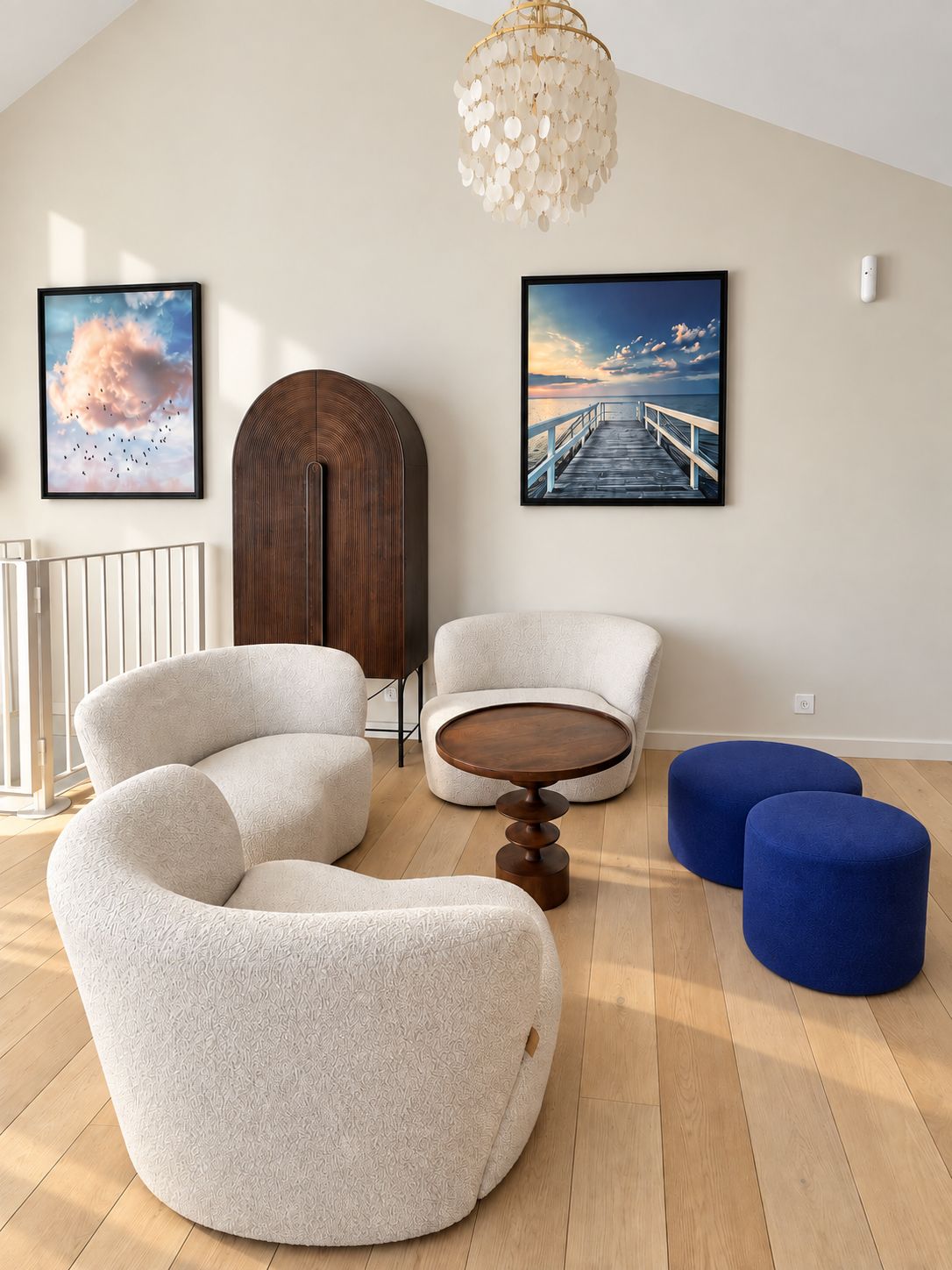 A modern living room with a wooden floor, a round coffee table, two sofas, and two ottomans. There are two framed pictures on the wall, a chandelier, and a white railing.