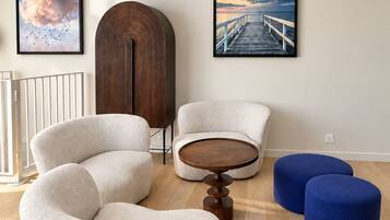 A modern living room with a wooden floor, a round coffee table, two sofas, and two ottomans. There are two framed pictures on the wall, a chandelier, and a white railing.