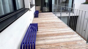 Una terraza de madera con bancos azules, un ventanal de vidrio que da a un espacio interior y una fachada de edificio moderna.