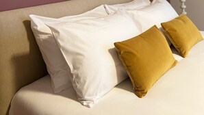 Una cama con almohadas blancas y mostaza, un espejo redondo en la pared y una lámpara de noche.