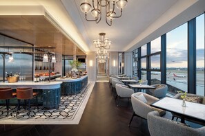 Um lobby moderno de hotel com área de bar, assentos e grandes janelas com vista para o aeroporto.