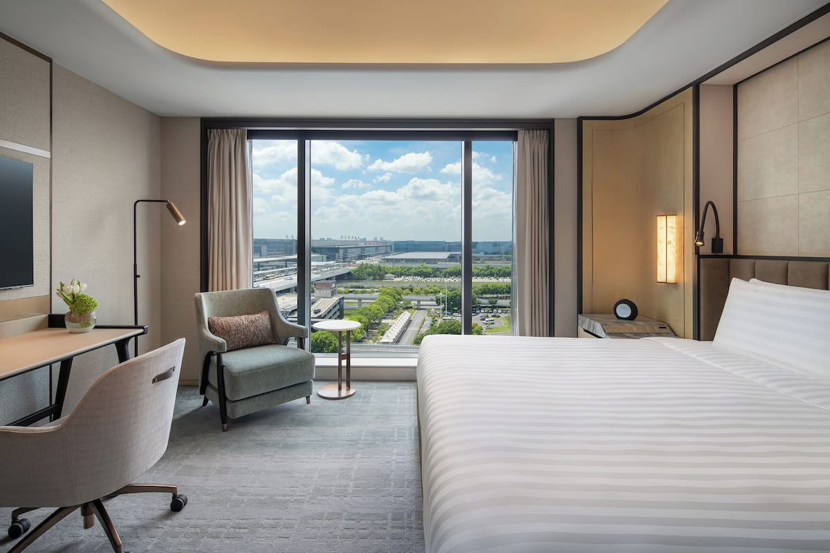 A modern hotel room with a large bed, a desk, a chair, and a view of the cityscape.