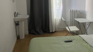 A small, simple room with a bed, a chair, a table, a sink, and a window with curtains. - Le Palmier (Saint-Denis)