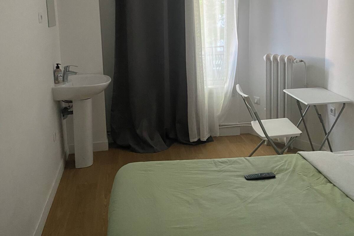 A small, simple room with a bed, a chair, a table, a sink, and a window with curtains.