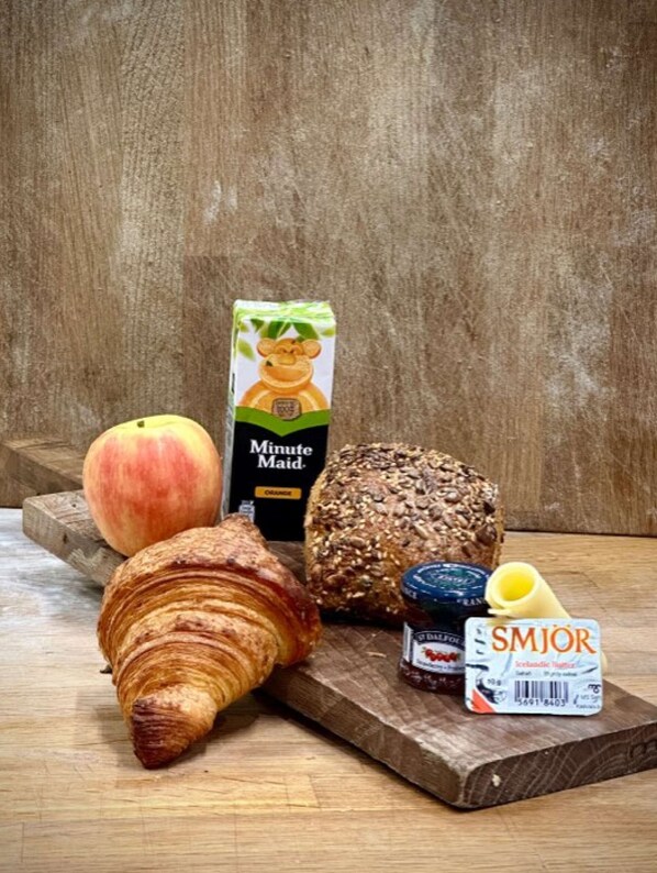 A wooden table with a croissant, a peach, a loaf of bread, a jar of jam, a carton of juice, and a bottle of cream.