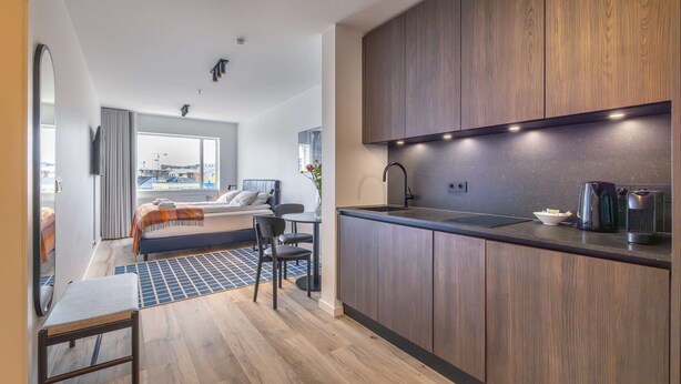 Una cocina moderna con armarios de madera, encimera negra y un lavamanos integrado. Hay una zona de comedor con mesa y sillas, y vistas a la ciudad a través de amplias ventanas.