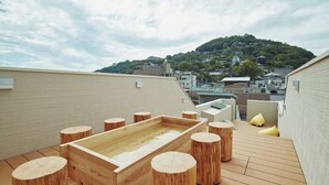A wooden deck with a rectangular wooden tub, surrounded by wooden stools, overlooking a cityscape with buildings and a hill. - MitO Onomichi (Onomichi)