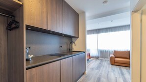 A modern kitchen with wooden cabinets and a black countertop, leading to a living area with a brown leather sofa and a large window. - Hlemmur Suites - Large Deluxe Studio (Reykjavík)