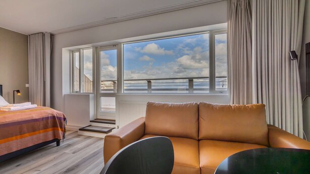 A hotel room with a bed, a sofa, a window with a view of the sky, and a curtain. - Hlemmur Suites - Large Deluxe Studio (Reykjavík)