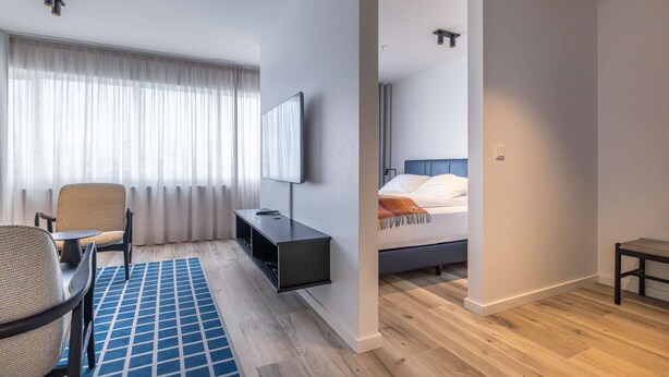 A hotel room with a bed, a desk, a chair, a TV, and a large window with curtains. - Superior one bedroom apartment (Reykjavík)
