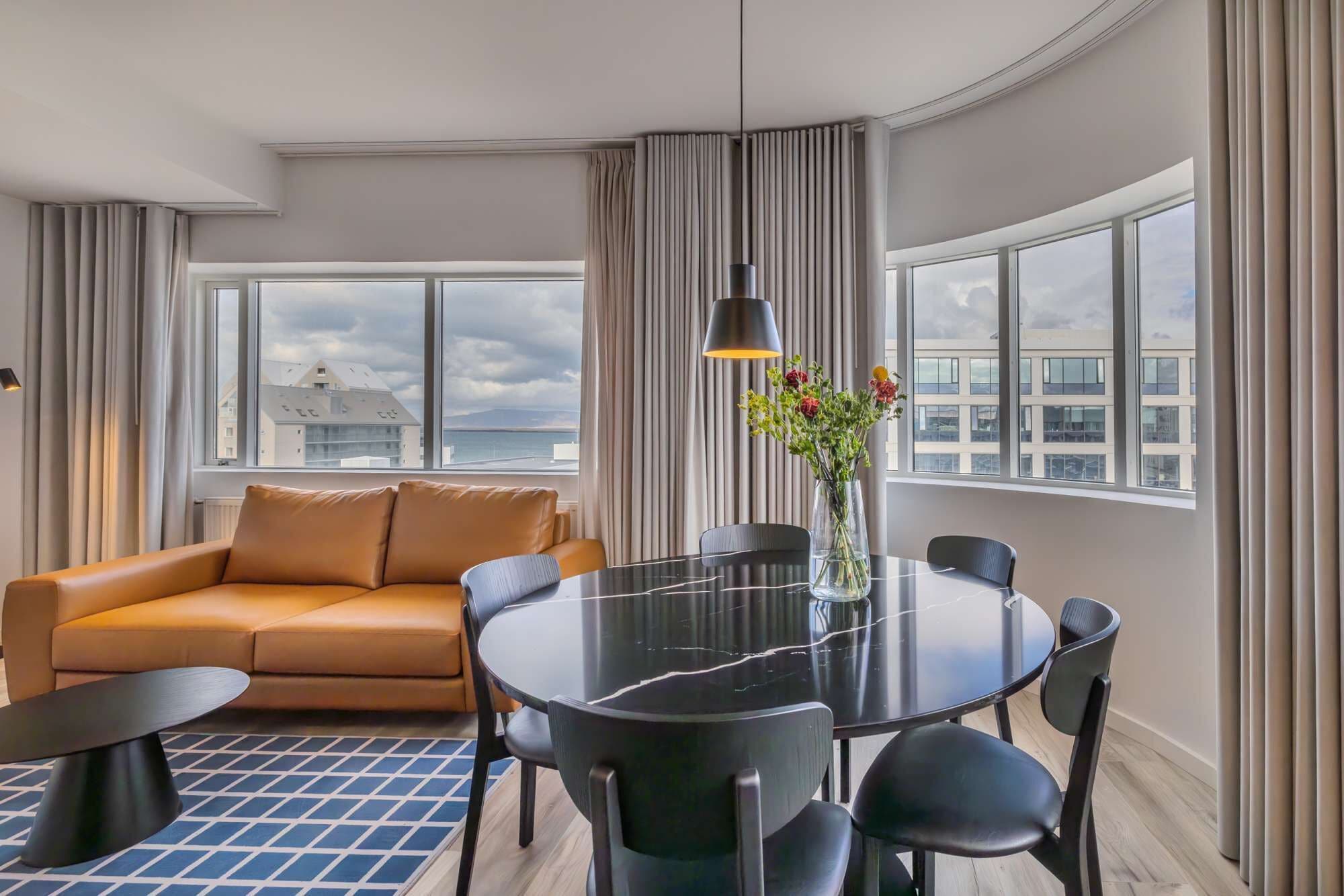 A modern hotel room with a leather sofa, a round dining table with chairs, a vase with flowers, and large windows with a city view.