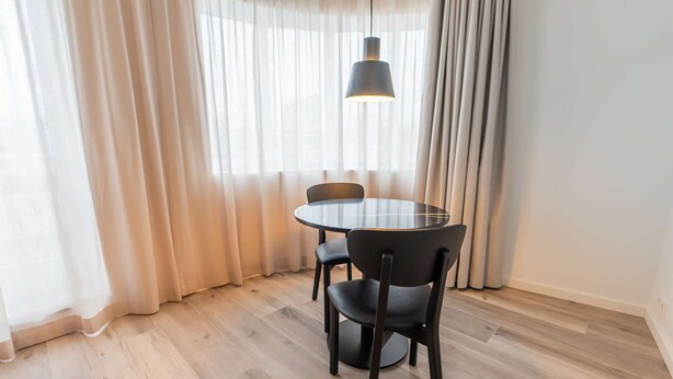 Un comedor con una mesa redonda y cuatro sillas, una lámpara colgante y cortinas traslúcidas.