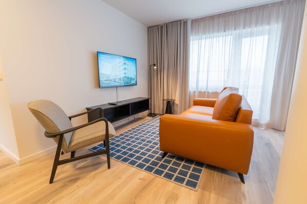 A modern hotel room with a flat-screen TV mounted on the wall, a sofa, a chair, and a small side table.