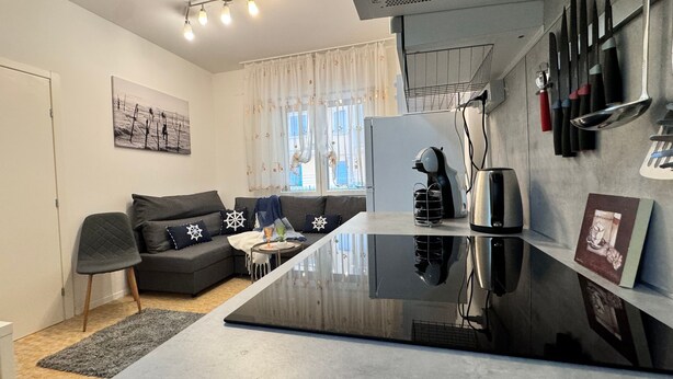 A modern kitchen with a black countertop, a built-in stove, and a coffee machine. There is a grey sofa with star-patterned cushions and a window with curtains. - Casa Angela by Ca-sa Check (Chioggia)