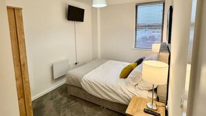 Une petite chambre à coucher moderne, avec un lit simple, une table de chevet avec une lampe, un téléviseur fixé au mur et une fenêtre avec des stores.