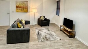 Un salon moderne avec un canapé gris, un fauteuil noir, un téléviseur à écran plat posé sur un meuble en bois, un tapis à motifs et un tableau accroché au mur.