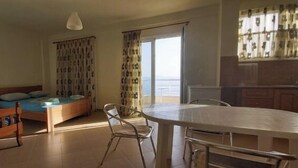 A room with a bed, a dining table, and a chair. There is a window with curtains, a ceiling light, and a view of the sea. - Magic Ionian Apartments & Rooms (Himarë)