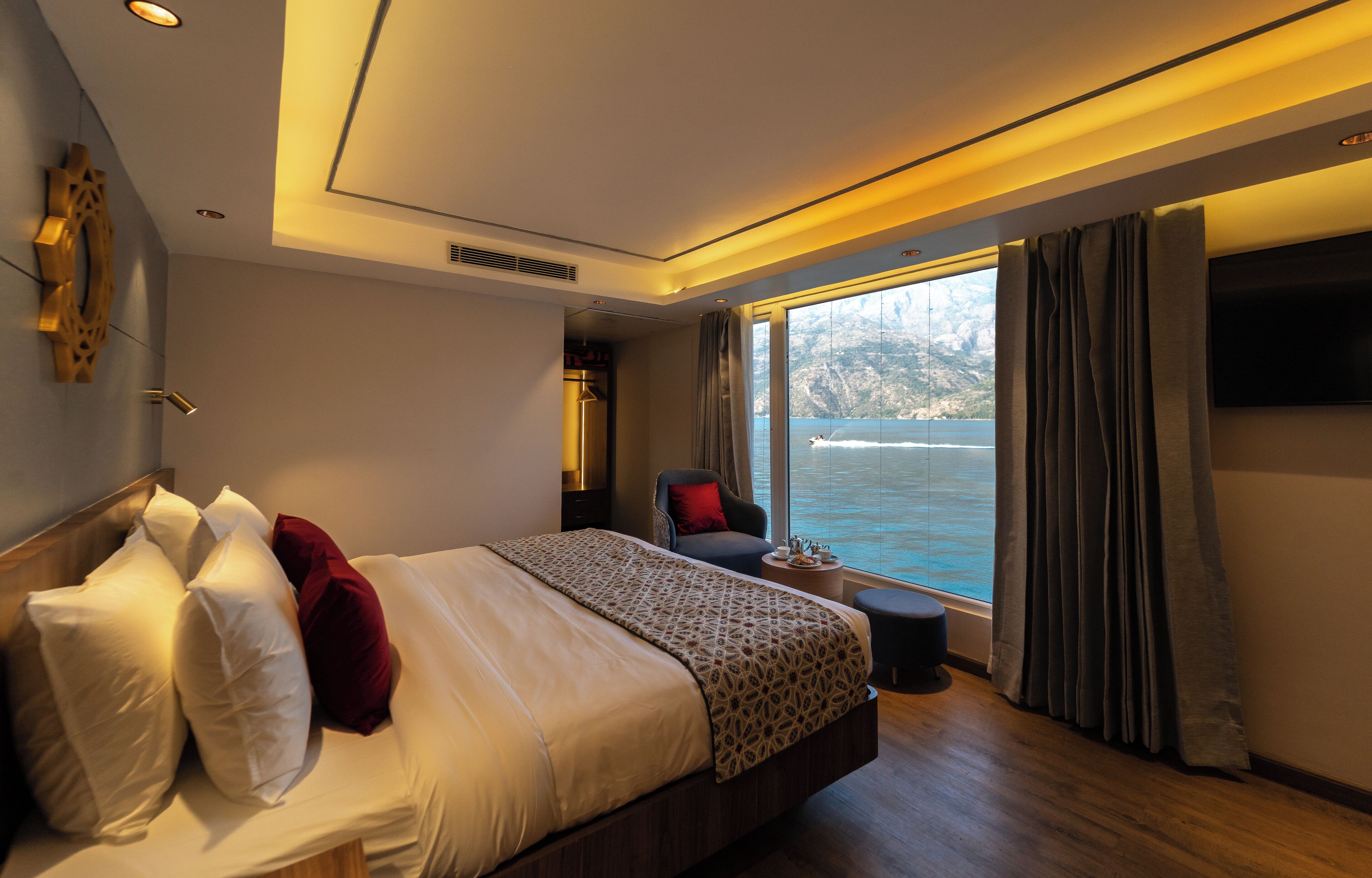 A modern hotel room with a large bed, a chair, a TV, and a view of the sea.