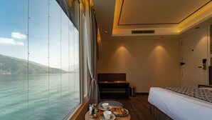 Une chambre d’hôtel dotée d’une grande fenêtre donnant sur un plan d’eau et des collines, d’un lit, d’une table de chevet avec une assiette de nourriture et des tasses, ainsi que d’un petit coin salon avec un banc.