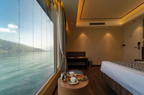 A hotel room with a large window offering a view of a body of water and hills, a bed, a bedside table with a plate of food and cups, and a small seating area with a bench. - Divine Lake Cruise (Tehri)