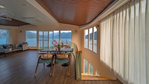 Un vaste espace de vie doté d’un plafond en bois, de grandes fenêtres offrant une vue sur la mer, et d’un coin repas avec des chaises et une table en osier.