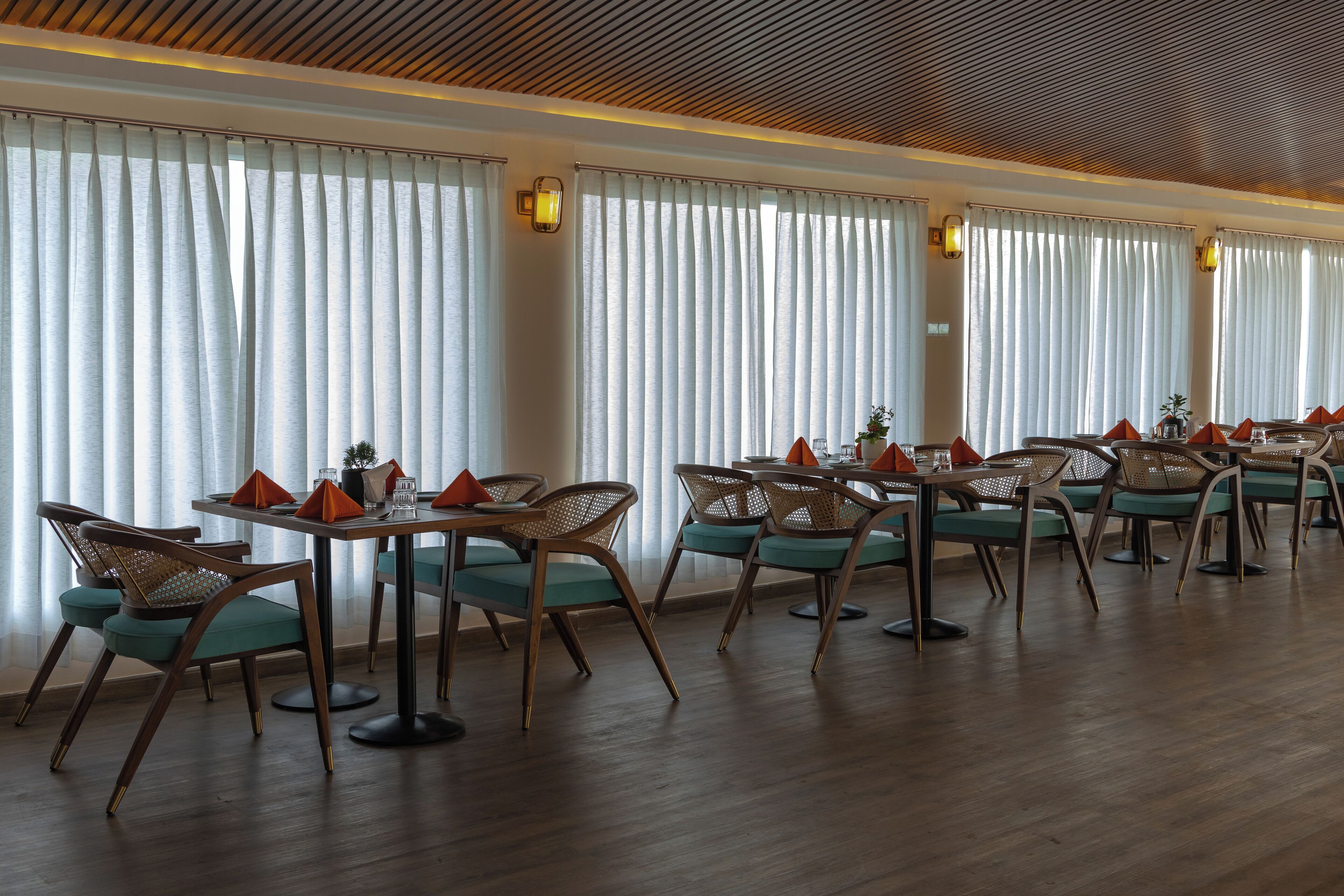 A restaurant with multiple tables and chairs, large windows with curtains, and a wooden ceiling.