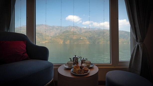 Une pièce avec vue sur un lac et des montagnes, une table avec un service à thé et un canapé agrémenté d’un coussin rouge.