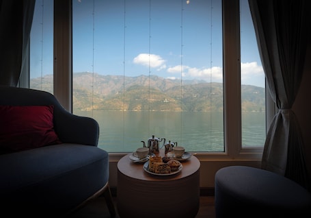 A room with a view of a lake and mountains, a table with tea set, and a sofa with a red pillow.