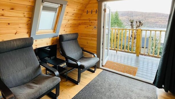 A cozy cabin interior with wooden walls, a balcony with a view of the mountains, and two grey chairs.